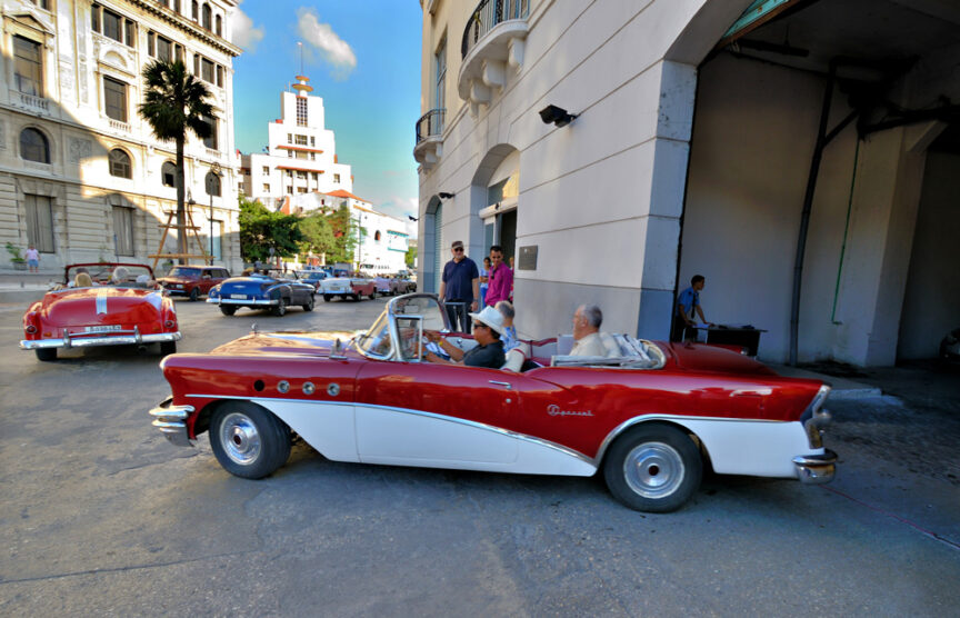 Classic red and white convertible with two passengers parked beside a white building; several other vintage cars, reminiscent of Cuba's streets, visible on street lined with people and old architecture.