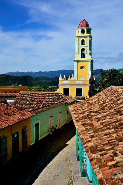 A tall, yellow bell tower stands prominently among colorful buildings with red-tiled roofs in a quaint Cuban town. Mountains and a partly cloudy sky provide a picturesque backdrop.