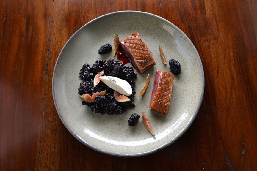 A plated gourmet dish featuring seared duck breast slices accompanied by blackberries, figs, and a dollop of cream on a brown ceramic plate set on a wooden table. This culinary trip offers a journey of the senses with every delicious bite.