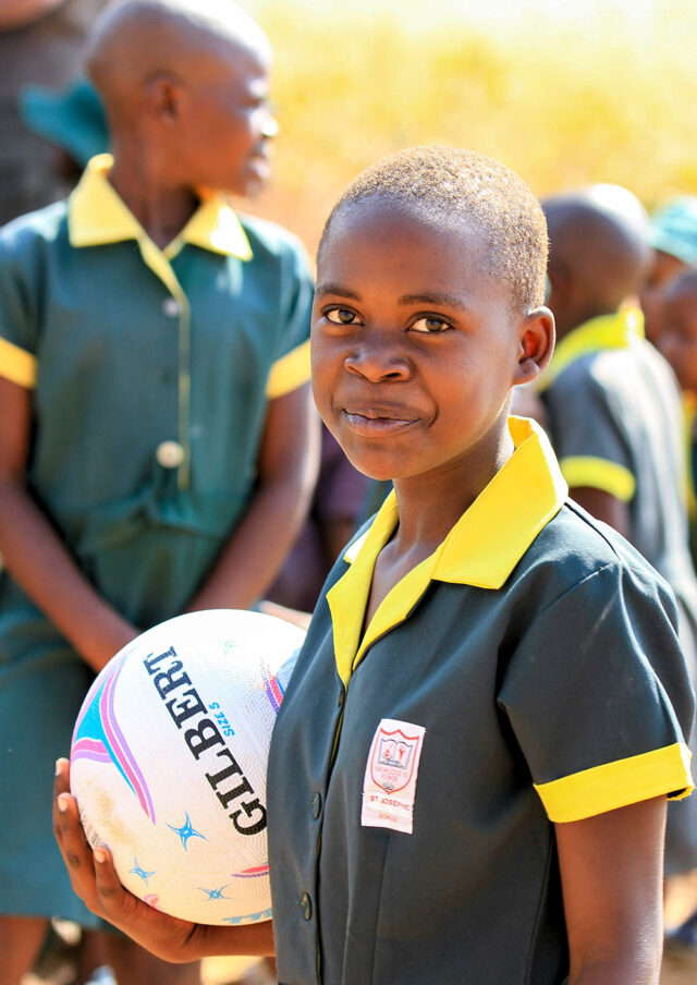 A smiling child in a school uniform holds a Gilbert netball, embodying pure joy. Other children in similar uniforms are visible in the background, creating a scene that feels like Heaven on Earth.