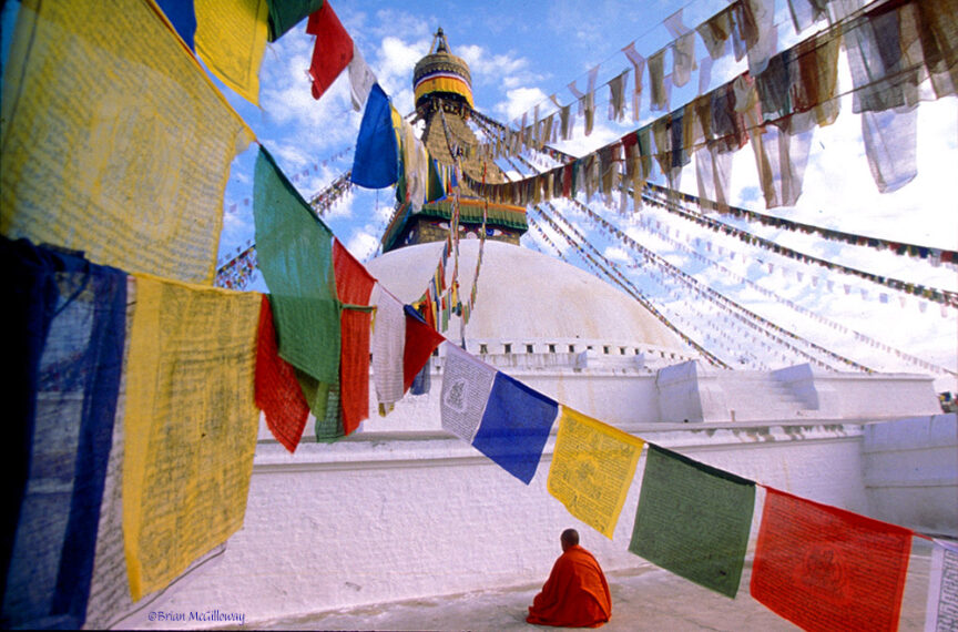 A lone person in an orange robe sits near a large white stupa adorned with colorful prayer flags against a blue sky, embodying the spirit of exploration in Nepal.