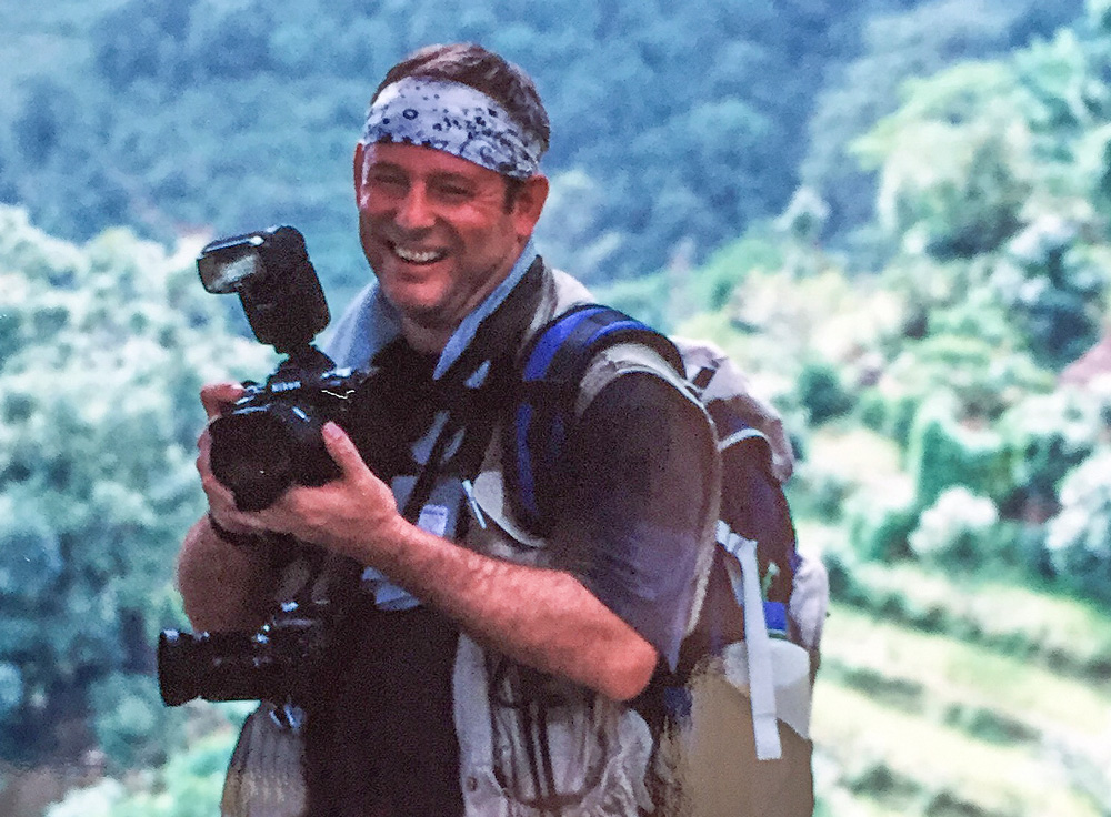 A person wearing a bandana and a backpack is smiling while holding a camera with a flash attachment, perhaps capturing moments for a family photobook. The background features a blurred green, forested landscape, reminiscent of Nepal's breathtaking scenery.