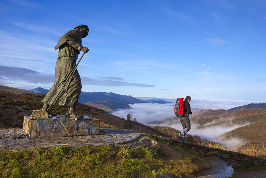A hiker with a red backpack walks on a historic path, passing beside a stone statue holding a staff, with fog-covered hills and a blue sky in the background.