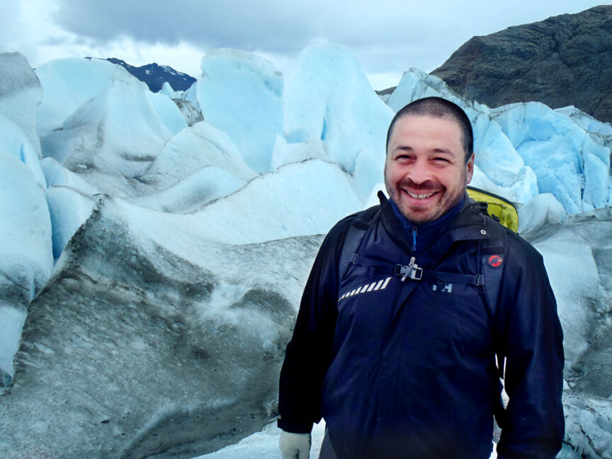Carlos Leon, in a black jacket, stands smiling in front of blue and white glacier formations under a cloudy sky for his personal profile.