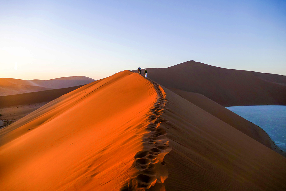 A group of tourists walking along the ridge of a sand dune in Namibia's desert landscape during sunset, with other dunes visible in the background.