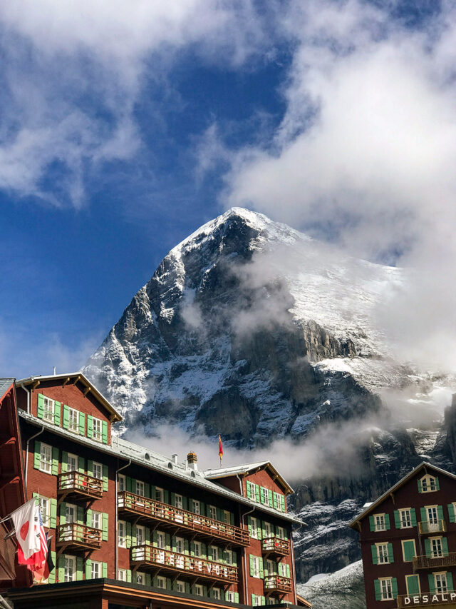 Red building with green shutters and balconies in front of a snow-covered mountain peak partially obscured by clouds. A Swiss flag proudly waves on the building, a common sight for those hiking along the Via Alpina in beautiful Switzerland.