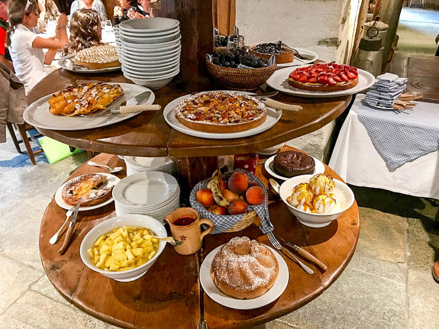 A multi-tiered dessert stand displays various pastries, cakes, and bowls of cut fruit, reminiscent of a sweet reward after hiking the Tour du Mont Blanc. In the background, people are dining at a restaurant table.