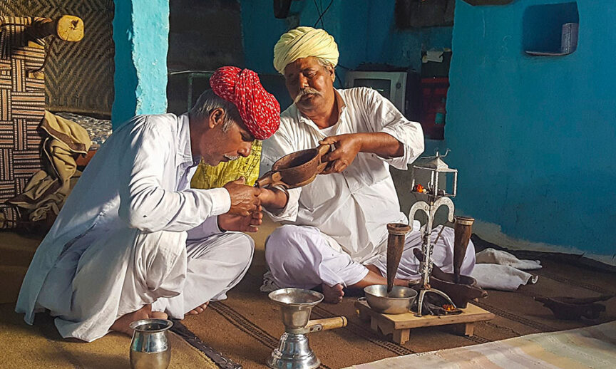 Two men in traditional Rajasthani attire engage in a ritual ceremony; one pours liquid from a bowl into the other's hand inside a blue-painted room, with various vessels and a water dispenser nearby, capturing the wonders of Rajasthan's rich cultural heritage.