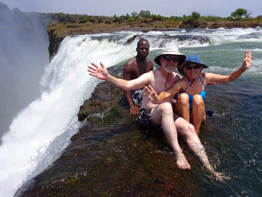 Three people in swim attire wave while sitting at the edge of the Smoke That Thunders waterfall, with water cascading nearby.