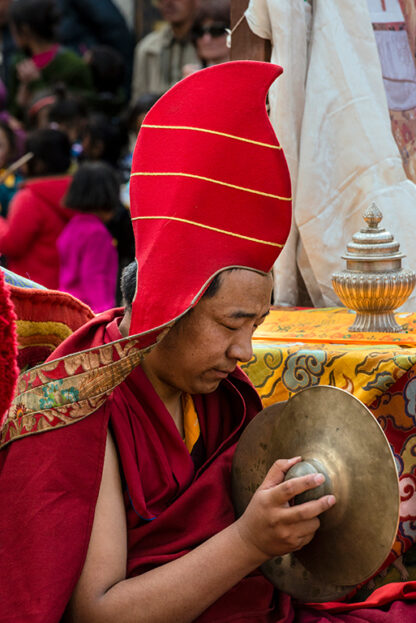 A seated individual in a red and gold hat is holding a brass dish. The person appears focused, with a colorful crowd and ornate objects in the background, reminiscent of the vibrant scenes at Nepal's Tiji Festival.