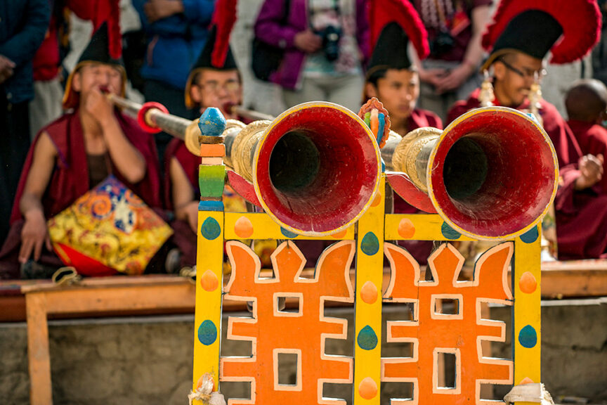 A close-up of two large, decorated horns with red interiors positioned on a colorful wooden stand during the Tiji Festival in Nepal, with people in traditional attire seated in the background.