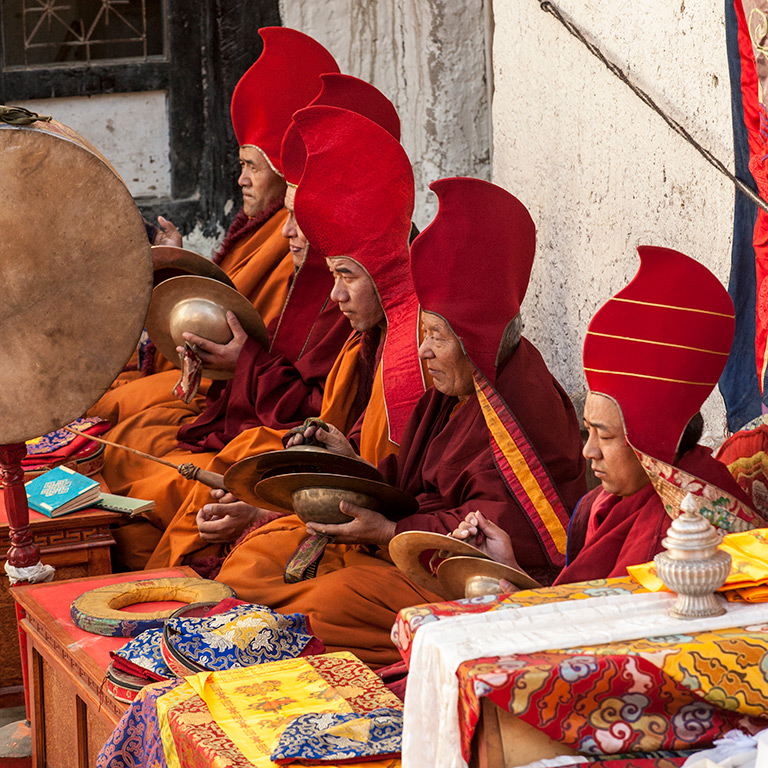 A group of monks in red hats and orange robes sit in a row performing a ritual with cymbals and a drum during the Tiji Festival in Nepal. Various colorful fabrics and objects are placed in front of them.