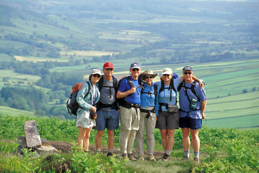 Six hikers with backpacks stand in a line, smiling at the camera, with a scenic green hilly landscape in the background. These adventurers savor the joy of traversing historic paths that countless others have walked before them.