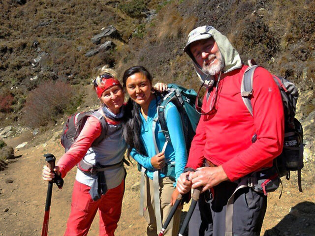 Three hikers in bright clothing, with backpacks and trekking poles, pose together on a rocky trail in a mountainous area under clear skies, their spirits lifted by the serene beauty and the silent echo of "Om Mani Padme Hum" in the peaceful surroundings.