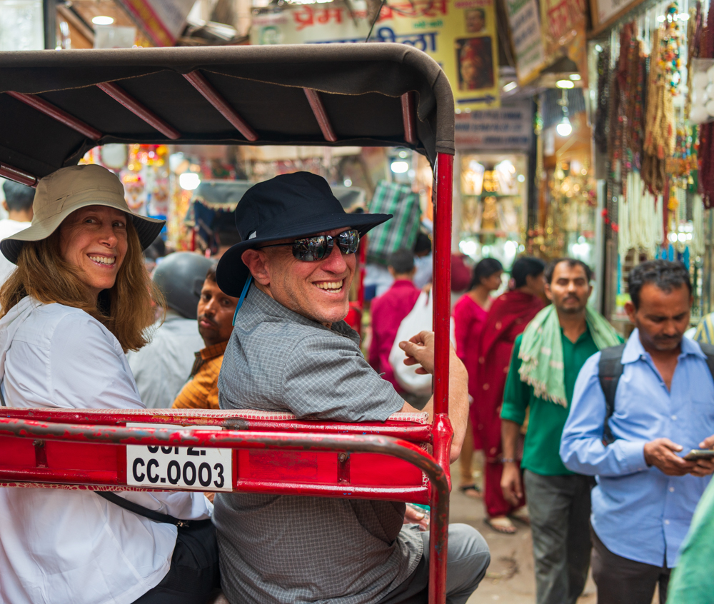 Two tourists in hats sit in a red rickshaw in a bustling market street of Northern India, surrounded by vendors and pedestrians. The man, wearing sunglasses, looks back and smiles while the woman looks forward and smiles, savoring their cultural journey.