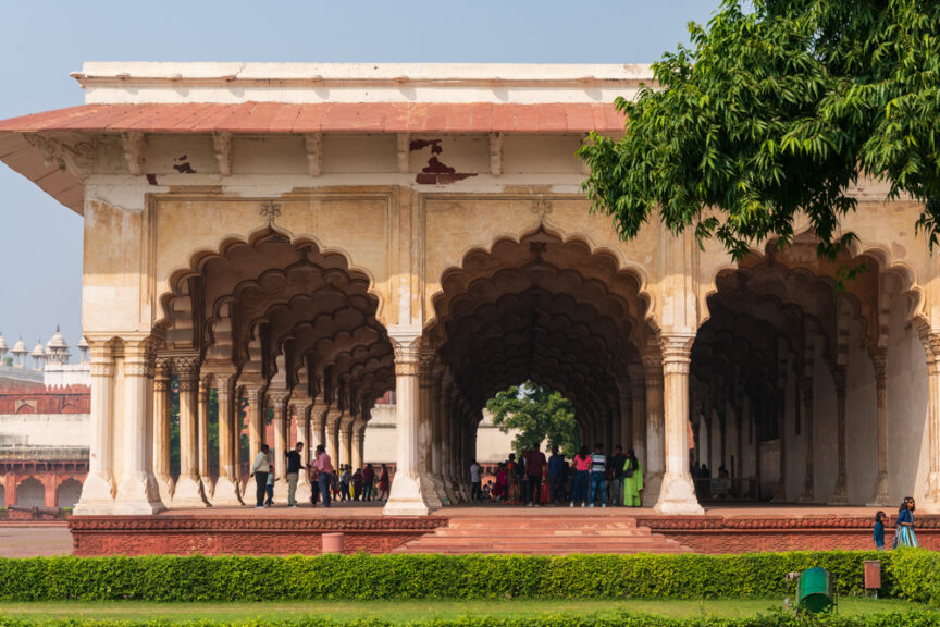 A group of people gathers under the arches of a historical pavilion with intricate designs, surrounded by a green lawn and a tree in the foreground, embarking on a cultural journey through Northern India.