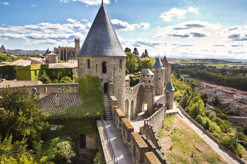 A historic stone castle with conical towers and ivy-covered walls is situated on a hill overlooking a village and lush countryside under a clear sky with scattered clouds, standing as one of the most picturesque citadels in the region.