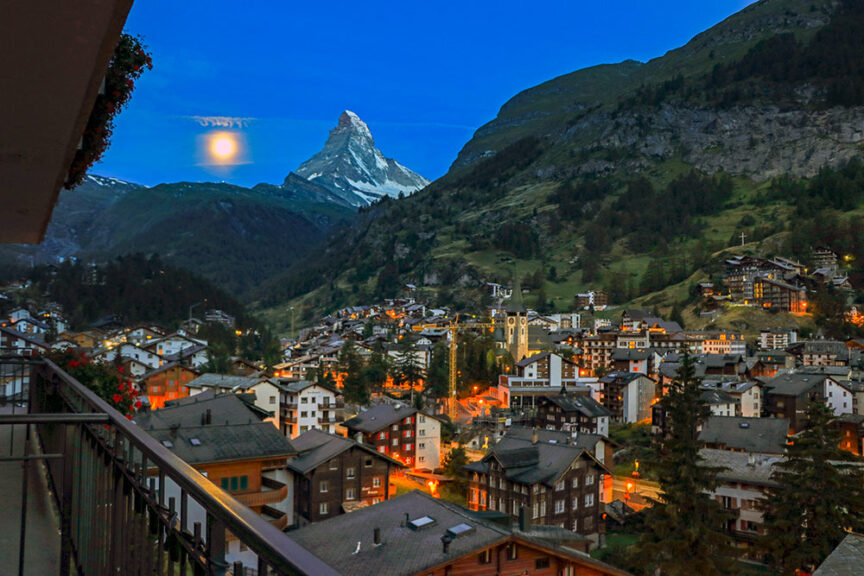 A view of a town nestled in a valley with the Matterhorn mountain in the background at dusk, illuminated by streetlights and a full moon in the sky, invites adventurers to dream of hiking trails that traverse through five countries.