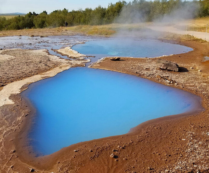 Two geothermal hot springs with vivid blue water surrounded by brown and rocky terrain, with steam rising and greenery in the background, make for an ideal stop during hikes in Iceland. Imagine watching the Northern Lights dance above as you soak in these natural wonders.