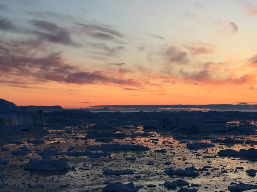 Frozen sea with scattered icebergs at sunset, featuring calm water reflecting the orange and pink hues of the sky, illuminated by soft light.