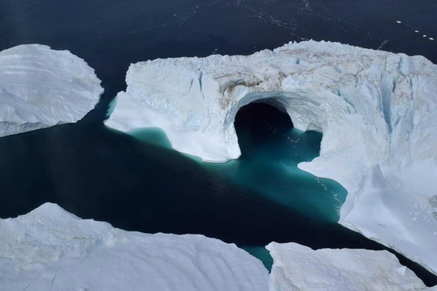 Aerial view of a large iceberg with an arch-shaped formation, its icy peaks glistening under the light, over dark blue water in a polar region.