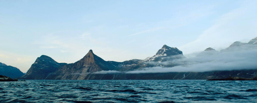 A rugged mountain range with icy peaks rises from the edge of a calm body of water, with a low layer of clouds hugging the mountains and a clear blue sky overhead.