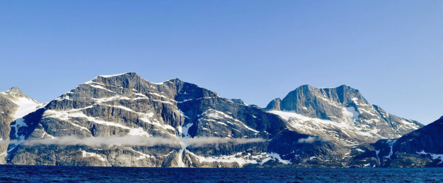 A panoramic view of snow-capped peaks under a clear blue sky, with light reflecting off a body of water in the foreground.