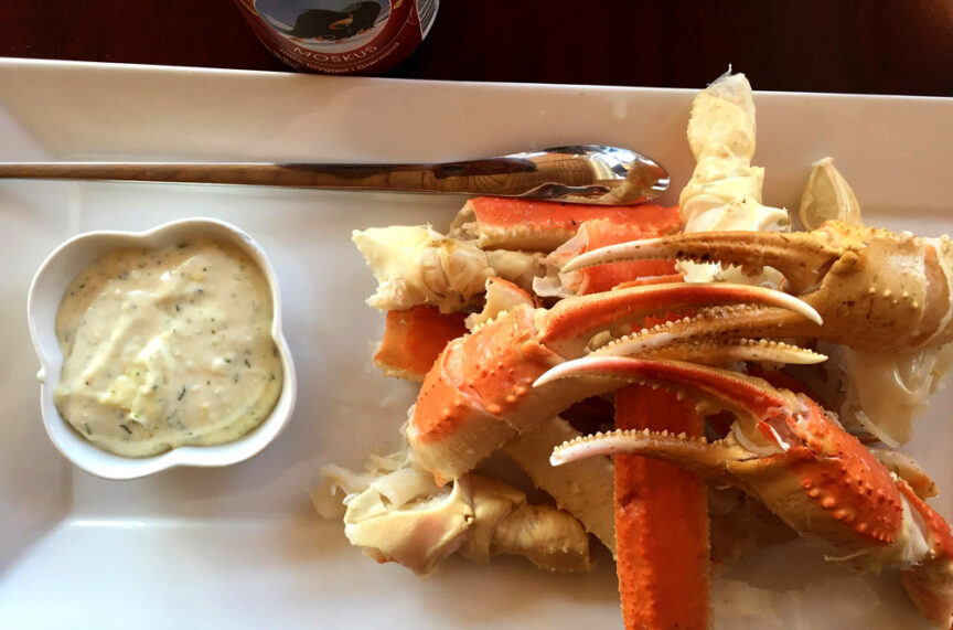 A white plate with crab legs next to a bowl of dipping sauce, accompanied by a spoon, all glistening under the light.