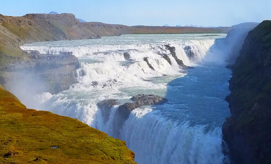 A wide, powerful waterfall cascades over a rocky cliff into a deep river gorge, surrounded by grass-covered plateaus under a clear blue sky, offering an idyllic spot for hikes in Iceland. Imagine witnessing the Northern Lights dancing overhead on a crisp evening here.