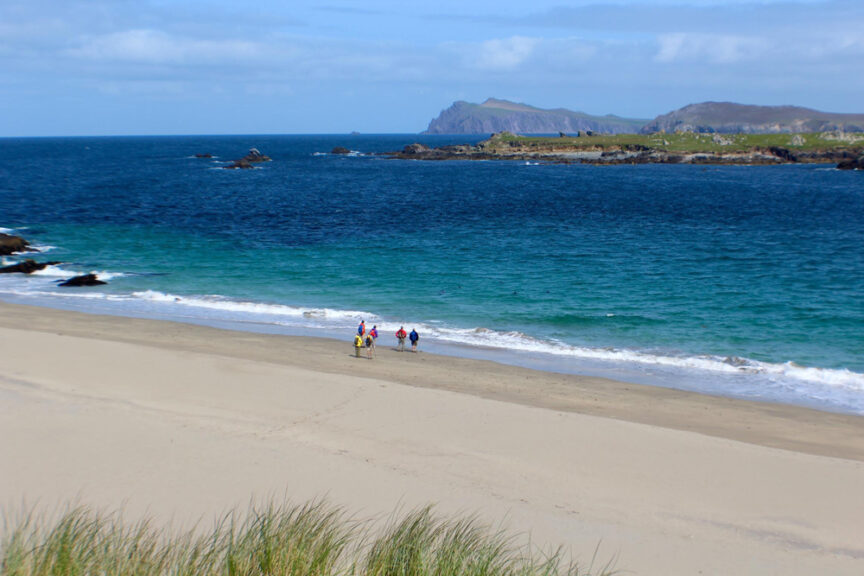 A beach with clear blue-green water, four people walking along the shore, and hilly islands in the background under a partly cloudy sky offers a scene that perfectly captures a sense of Céad Míle Fáilte.