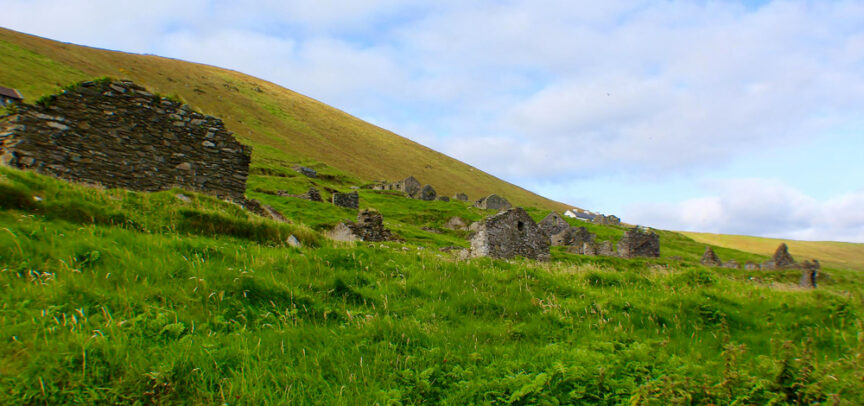 Ruins of stone buildings scattered across a green hillside under a partly cloudy sky, offering a Céad Míle Fáilte to all who visit.