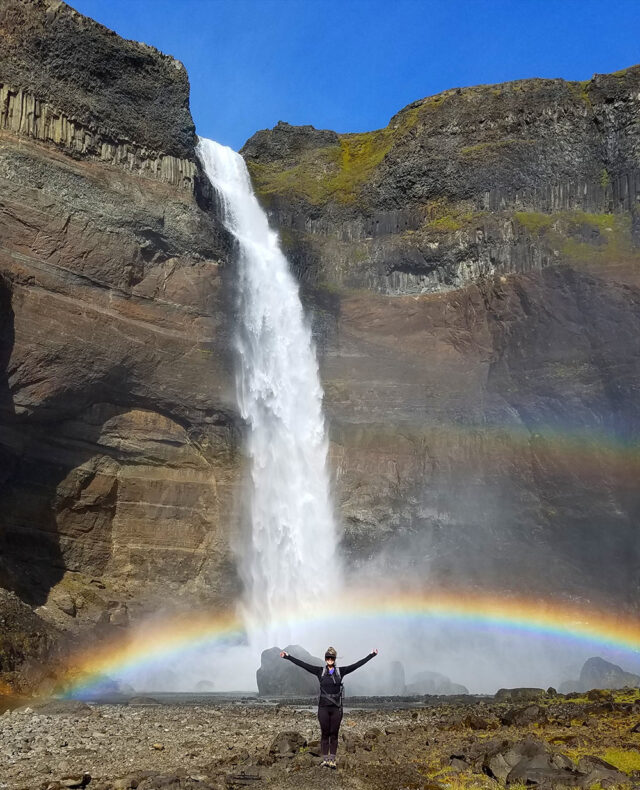A person stands with arms raised in front of a tall waterfall cascading down a rocky cliff during one of the famed Iceland hikes, with a rainbow visible at the waterfall's base.