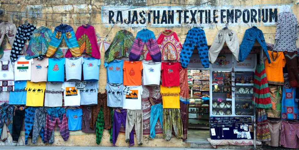 A storefront named "Rajasthan Textile Emporium" displaying vibrant fabrics, clothes, and garments. The front wall is adorned with hanging colorful textiles and shirts, reflecting the diverse beauty of this country of contrasts.