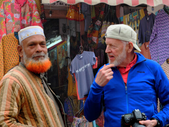 Two men, embodying the contrasts of their country, stand in front of a colorful market stall. One has a dyed orange beard and wears traditional attire, while the other wears a blue jacket and cap, holding a camera and gesturing animatedly.