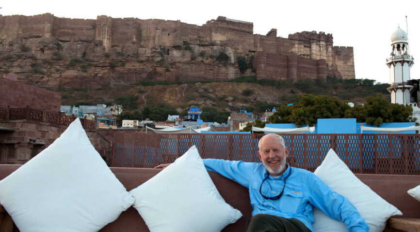 A man with a white beard sits on an outdoor couch with white pillows, smiling. Behind him, the striking contrasts of blue buildings and a large ancient fort on a hill paint the picturesque charm of this country.