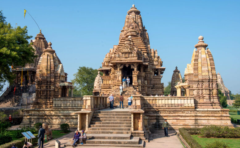 Tourists visit intricately carved stone temples with towering spires at the Khajuraho Group of Monuments in India, marveling at the contrasts between ancient artistry and modern life on a sunny day.