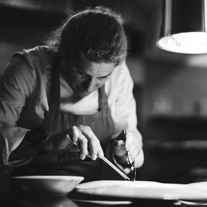 A chef in a kitchen, intensely focused on plating a dish with precision using tweezers, under the light of a hanging lamp, offering a delicious culinary journey of the senses.