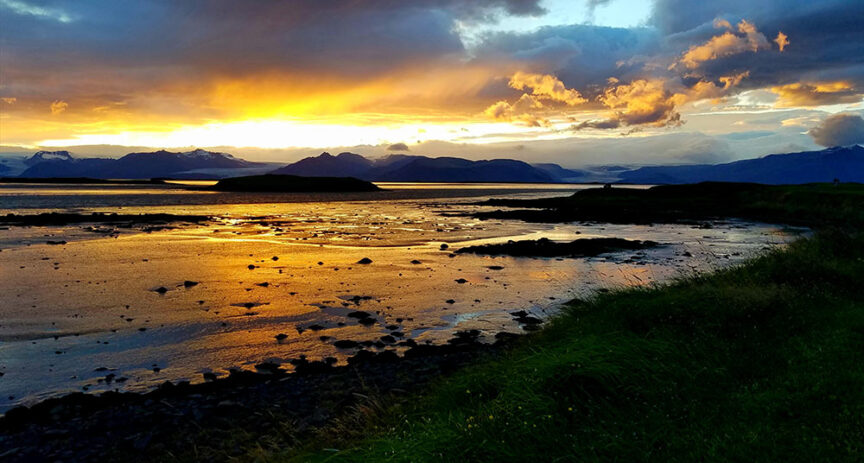 Sunset over a coastal landscape in Iceland with mountains in the background. The sky is partly cloudy with vibrant hues of orange, yellow, and blue reflecting on the water. Rocky shoreline and grassy foreground visible, ideal for scenic hikes under the magical glow of the Northern Lights.