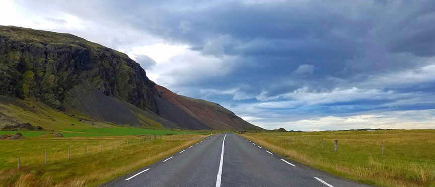 A straight road flanked by grassy fields stretches toward distant mountains under a cloudy sky, reminiscent of the scenic hikes in Iceland.