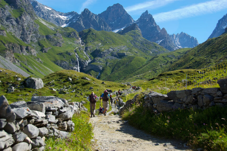 A group of hikers walk along a stone-walled path surrounded by green hills and Mediterranean rocky mountains under a clear blue sky.
