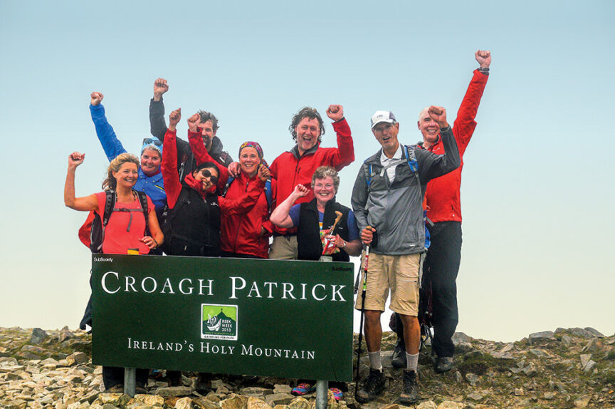 A group of hikers celebrates at the summit of Croagh Patrick, one of the world's iconic peaks, posing with raised arms behind a sign that reads "Croagh Patrick, Ireland’s Holy Mountain.