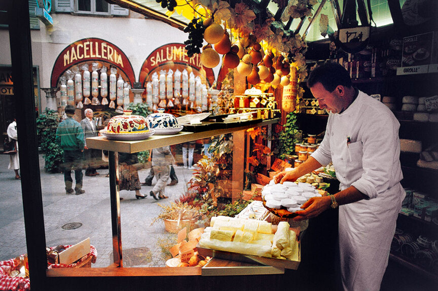 A shopkeeper arranges delicious cheese in a delicatessen, tantalizing the senses with an assortment of other food items on display. Through the window, a vibrant street scene with pedestrians and shops adds to the charm, making it a perfect spot for culinary trips.