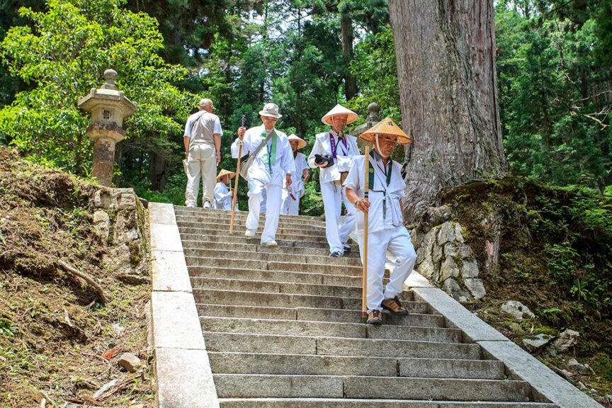 A group of people in traditional Japanese attire walk down stone steps in a forested area, holding walking sticks, as they embark on one of the historic paths traveled during pilgrimages.