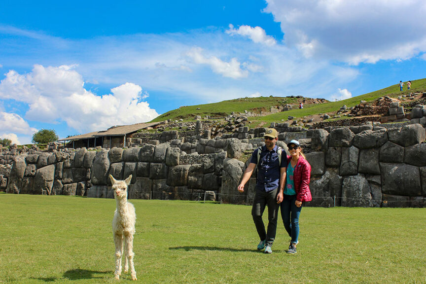 A couple walks together near ancient stone ruins, following in the footsteps of the Incas, with a llama standing in the grass in the foreground under a blue sky with scattered clouds.
