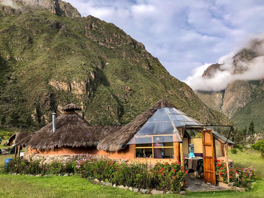 A round building with a thatched roof and partial glass dome stands amidst a lush, mountainous landscape, reminiscent of following the footsteps of the Incas. Flowers bloom around the building and clouds touch the mountain peaks in the background.