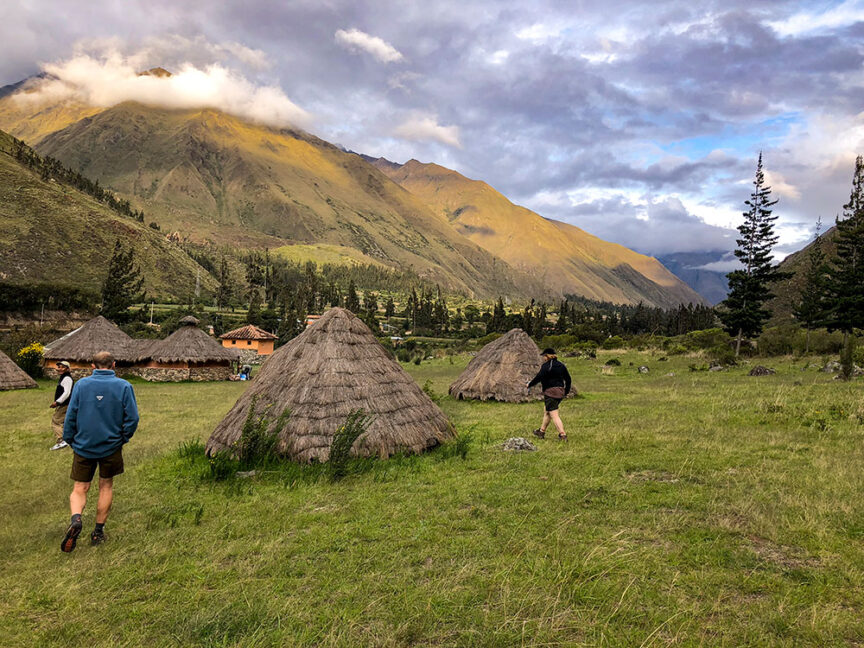 Three people, perhaps Incas, walk towards a settlement with thatched huts in a valley surrounded by mountains under a partly cloudy sky, their footsteps softly echoing through the serene landscape.
