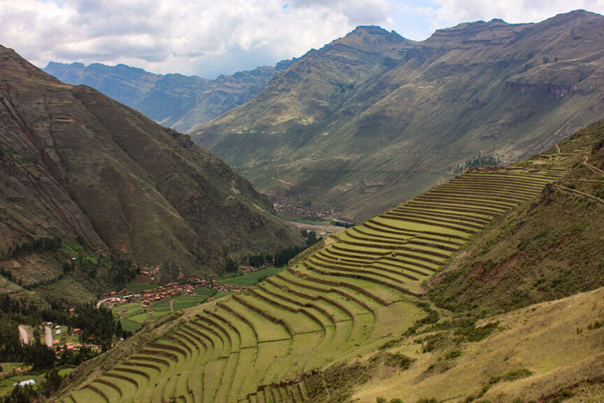 Terraced agricultural fields stretch across a steep mountainside, following in the footsteps of the Incas, with a valley and distant mountains under a cloudy sky.