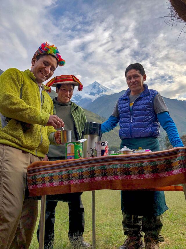 Three men are smiling and standing outdoors at a table with drinks, following in the footsteps of past adventurers. One man is holding a cup. Snow-capped mountains can be seen in the background.