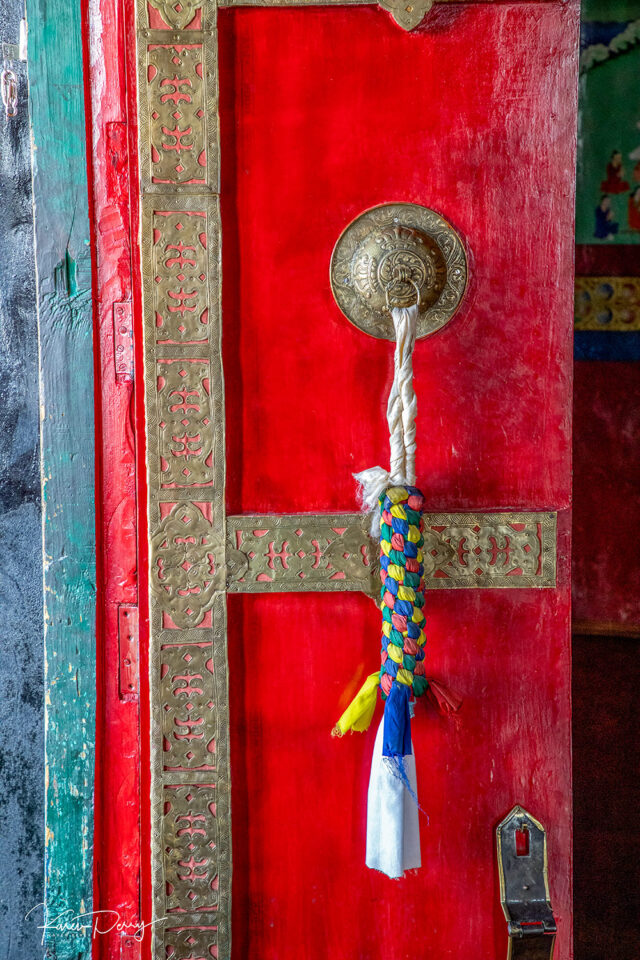 A vibrant red door with gold detailing and a brass handle, adorned with a colorful braided rope charm, stands as striking as the elusive beauty of snow leopards in their natural habitat.