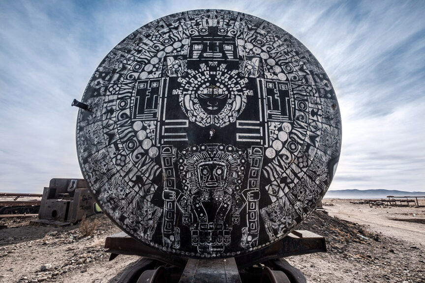 A photograph captures a large concrete circular structure with intricate black-and-white geometric designs, radiating surreal beauty as it sits in the desolate landscape of Salar de Uyuni under a cloudy sky.